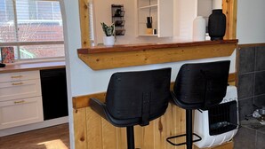 A kitchen with wooden accents, two black bar stools, a white refrigerator, and a window with blinds.