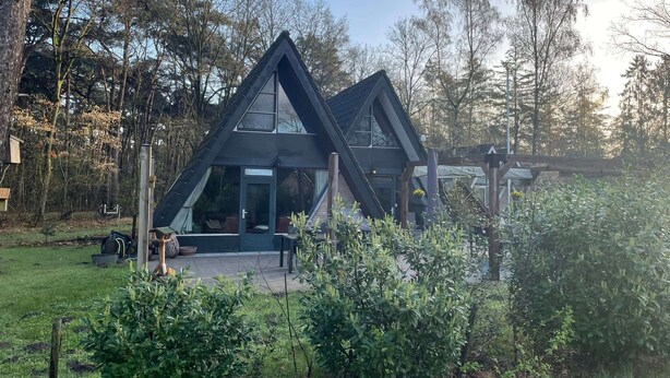 A modern, triangular-shaped house with large windows and a porch, surrounded by lush greenery and trees. - Bungalow in Limburg Near Nature Reserves (Stramproy)