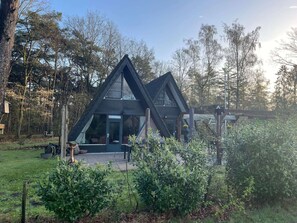 Ein modernes, dreieckiges Haus mit großen Fenstern und einer Veranda, umgeben von üppiger Bepflanzung und Bäumen.