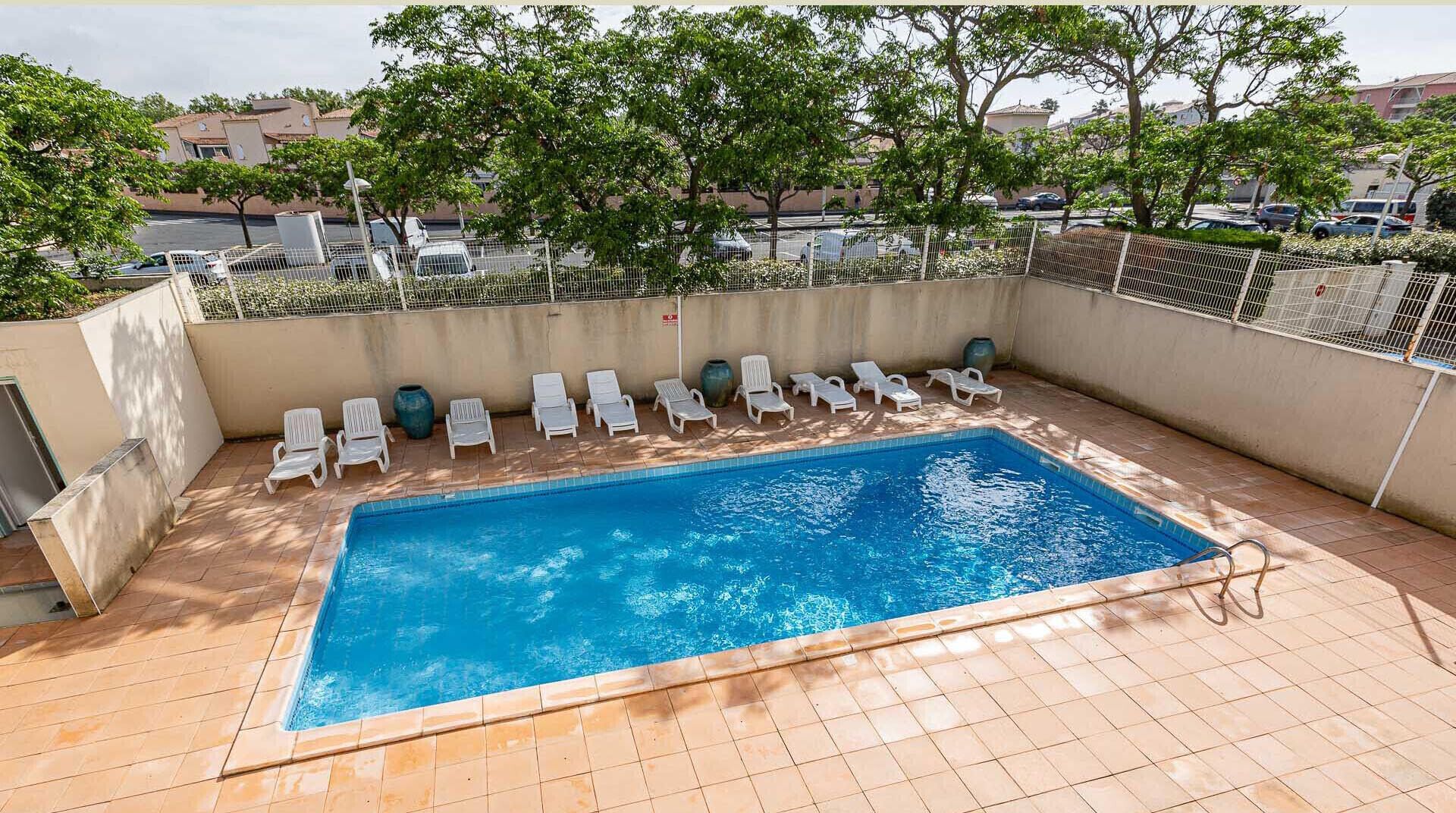 A rectangular swimming pool with a tiled deck, surrounded by white lounge chairs, and a view of buildings in the background.