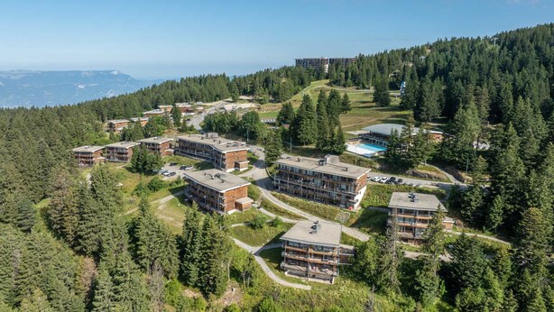 A high-rise hotel with multiple floors, surrounded by dense forest and a view of distant mountains. - Near the slopes, nice Appt for 6 pers (Chamrousse)