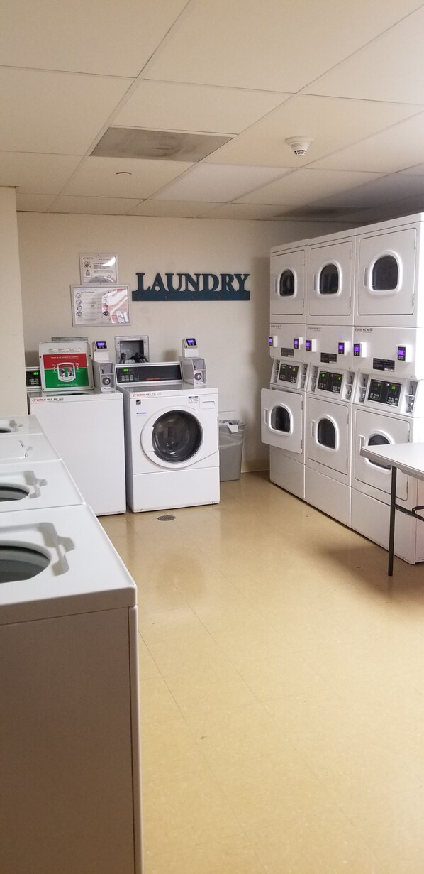 A laundry room with multiple washing machines and dryers, a wall-mounted sign reading "LAUNDRY", and a trash bin.