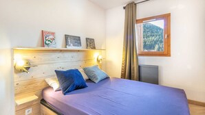 A bedroom with a wooden headboard, a bed with a purple bedspread, two pillows, a nightstand, a bedside table, a wall-mounted shelf with books, a wall-mounted light, a window with curtains, and a view of a forest. - Flat at the foot of the slopes with a balcony (Modane)