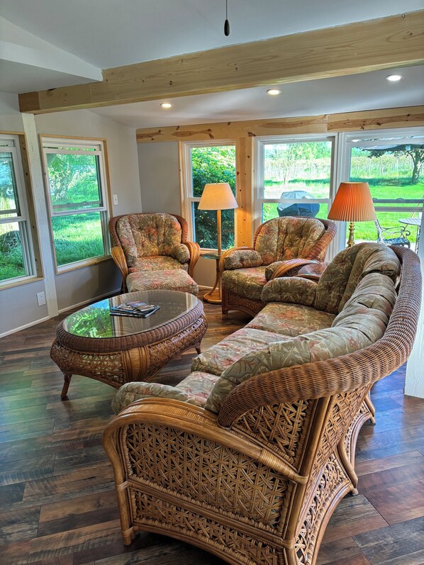 A spacious living room with wicker furniture, a glass coffee table, and large windows offering a view of the outdoors.