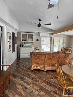 A spacious room with a herringbone wood floor, a wicker sofa, a dining table with chairs, and a ceiling fan.