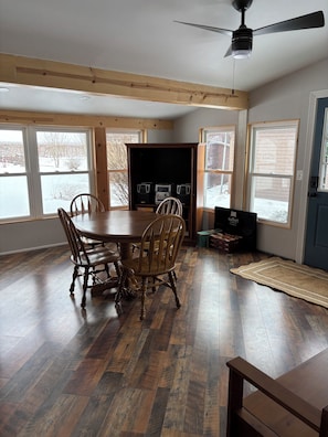 A dining area with a wooden table and chairs, large windows, and a ceiling fan.
