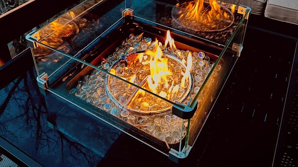 A glass-top fire pit with burning logs, surrounded by a wooden deck and a cushioned outdoor seating area.