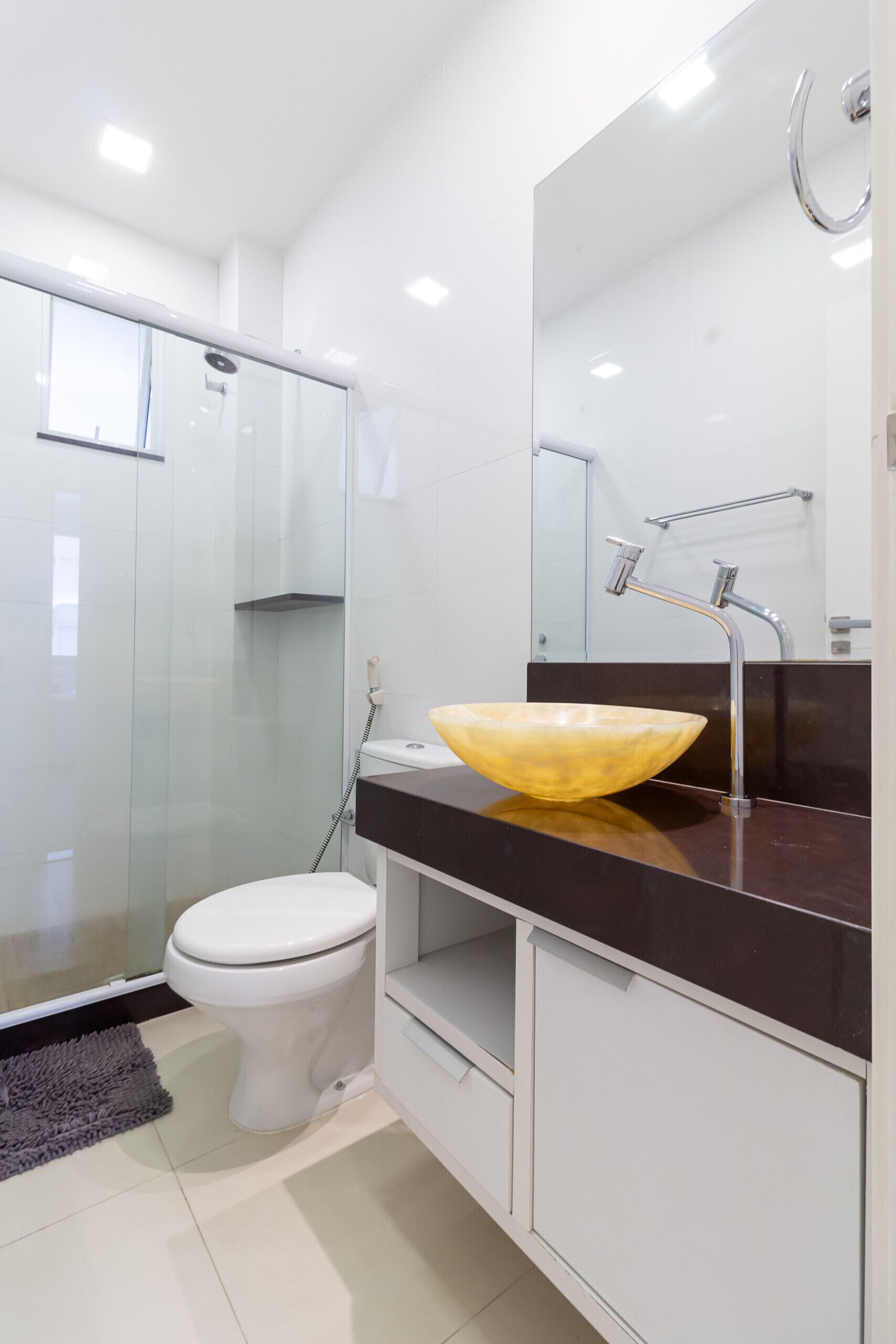 Une salle de bain moderne avec une douche à l’italienne en verre, des toilettes blanches, un grand miroir et un plan de toilette foncé surmonté d’un lavabo sur pied jaune.