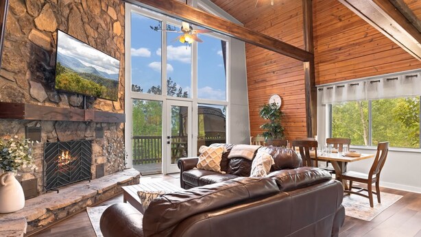 A stone fireplace, leather sofa, dining table, and large windows in a room with wooden beams. - Secluded Cabin for 10, Near Pkwy (Pigeon Forge)