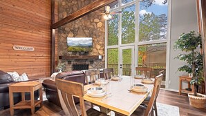 A cozy room with a stone wall, a wooden sofa, a dining table set for six, and a large window offering a view of trees. - Secluded Cabin for 10, Near Pkwy (Pigeon Forge)
