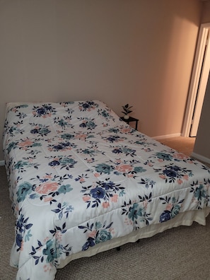 Uma cama com colcha florida, uma pequena mesa com uma planta e uma porta ao fundo.
