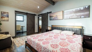 Une chambre avec un grand lit, un bureau et une chaise. Il y a une fenêtre et une porte qui donne sur une autre pièce.