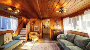 L’intérieur chaleureux d’un chalet avec des murs en bois, un canapé, un fauteuil, un tapis et un escalier menant à un espace cuisine.