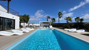 Una piscina con acqua cristallina blu, circondata da comode poltrone bianche, palme e architettura moderna.