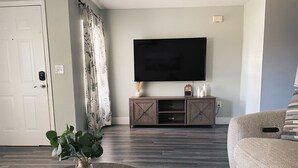 A living room with a dark wood coffee table, a beige sofa, a flat-screen TV on a wooden cabinet, and a door with a white frame.