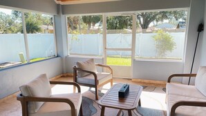 A sunlit patio with two white cushioned chairs, a small wooden table, and a view of a backyard through large windows.