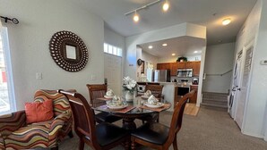 A dining area with a round table set for four, surrounded by wooden chairs. A colorful armchair and a small table with a vase and flowers are visible. A kitchen with modern appliances is seen in the background. - 4 Bedroom 3 Bathroom Villas At Seven Dwarfs 28-107 (Kissimmee)