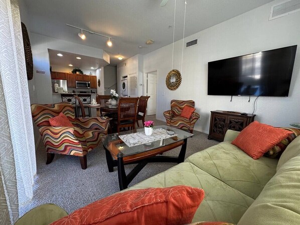 A living room with a green sofa, two patterned armchairs, a coffee table, and a flat-screen TV mounted on the wall. - 4 Bedroom 3 Bathroom Villas At Seven Dwarfs 28-107 (Kissimmee)