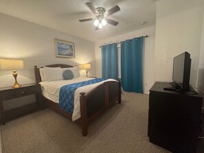 Un dormitorio con cama, lámparas de noche, ventilador de techo, televisor sobre un mueble y una ventana con cortinas.