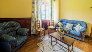 A living room with a blue sofa, two wooden chairs, a wooden coffee table, and a window with yellow curtains. - Charming cottage (Aldudes)