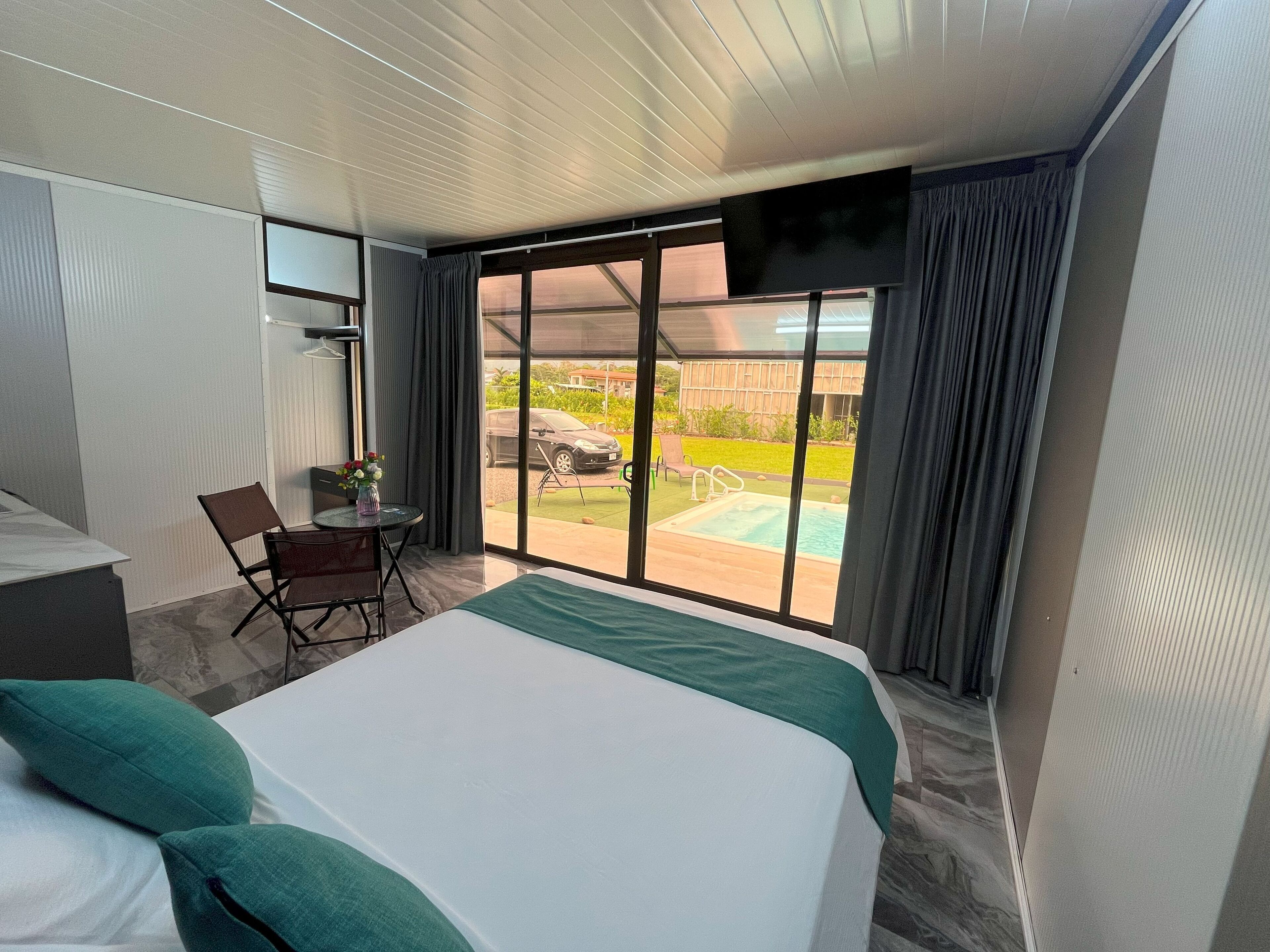 A hotel room with a bed, a desk, a chair, a small table with a vase, and a view of a swimming pool and outdoor area.