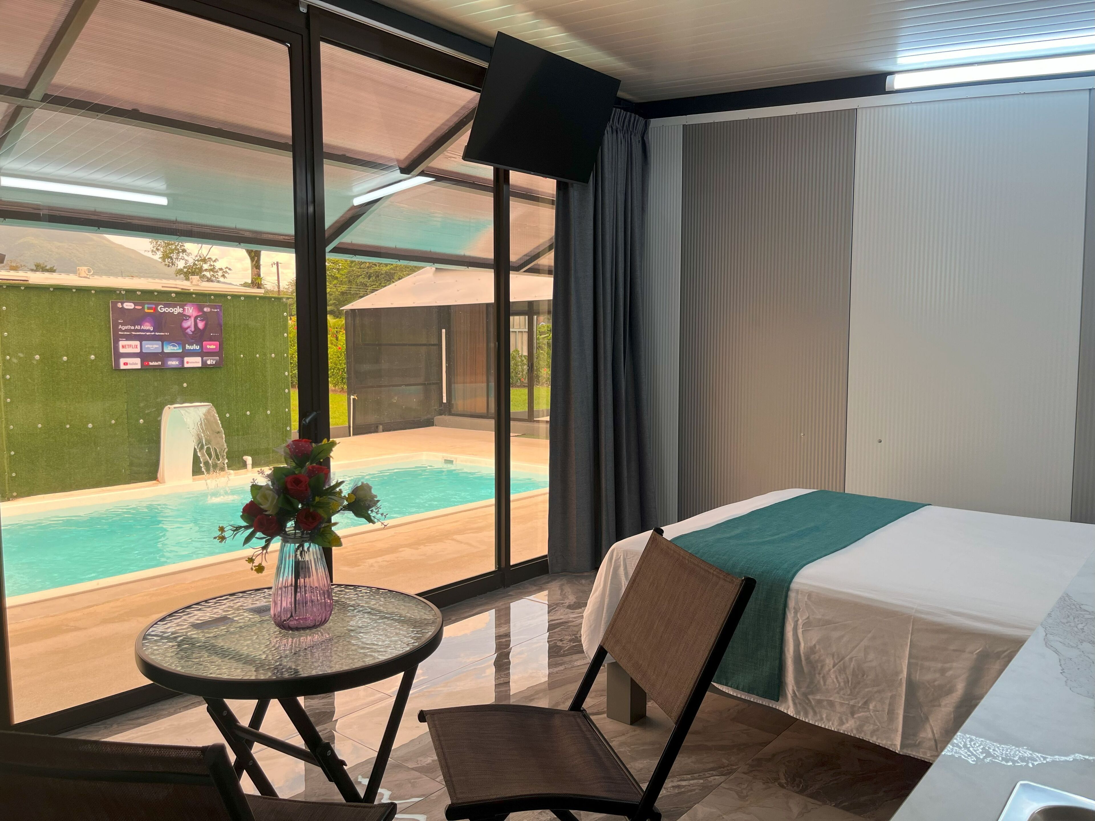 A hotel room with a bed, a table with a vase of flowers, a chair, a view of a swimming pool, and a large window with curtains.