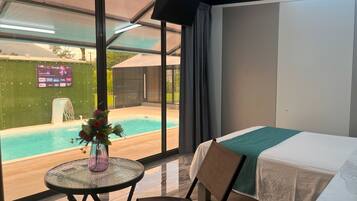 A hotel room with a bed, a table with a vase of flowers, a chair, a view of a swimming pool, and a large window with curtains.
