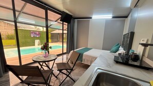 Quarto de hotel pequeno com uma cama, uma mesinha com cadeiras, pia e vista para uma piscina.