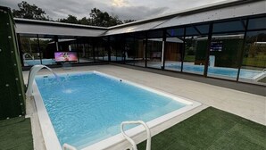 Piscina com estrutura de vidro, cercada por grama sintética e uma área coberta com televisão.