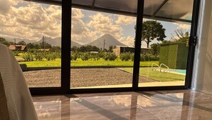 Quarto com uma grande janela que oferece vista para uma área ajardinada com piscina e uma montanha ao longe.