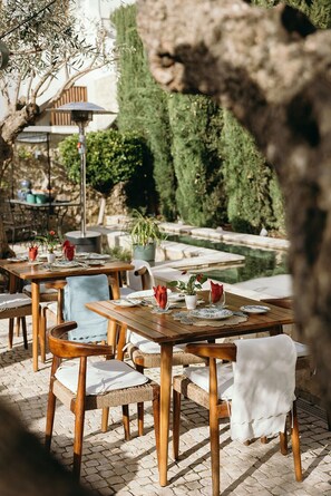 An outdoor dining area with wooden tables and chairs, a small pool, and greenery in the background. - Elevem Boliqueime  (Loulé)