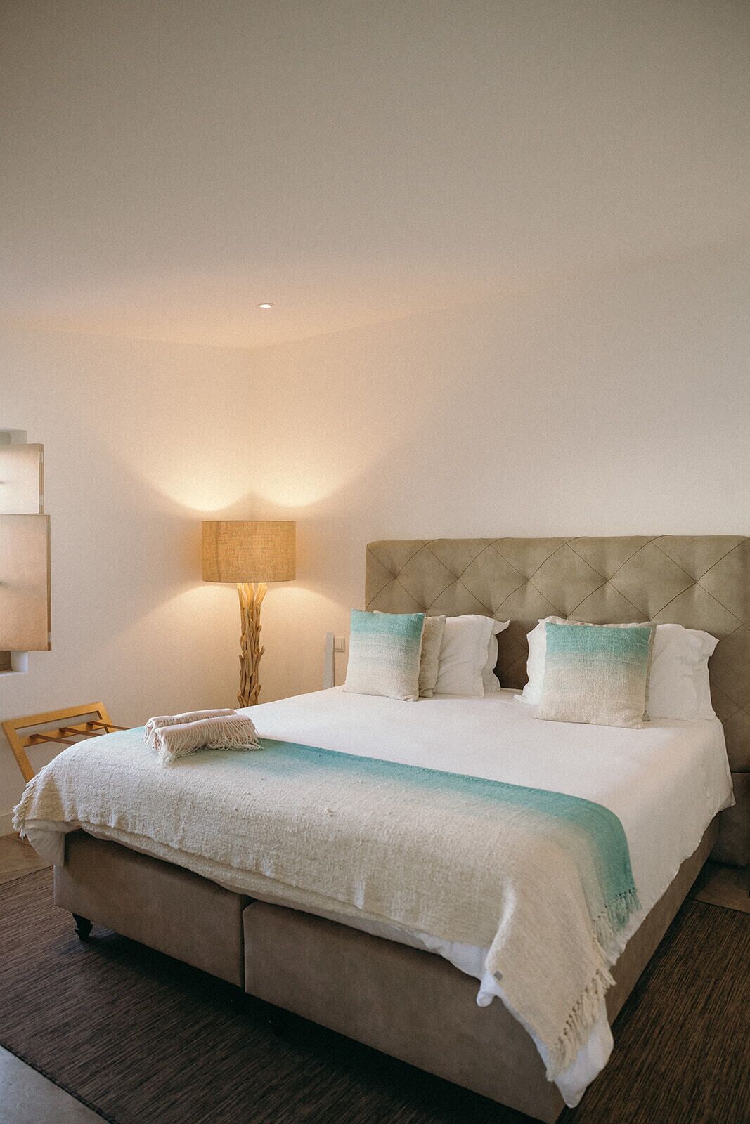 A neatly made bed with a beige headboard and white bedding, a wooden chair, and a floor lamp.