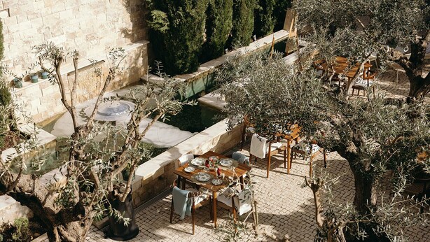 Un área de comedor al aire libre con suelo de piedra, olivos y una pared de piedra al fondo.
