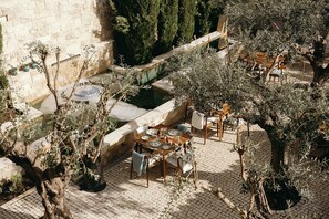 Un área de comedor al aire libre con suelo de piedra, olivos y una pared de piedra al fondo.