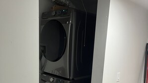 Un cuarto de lavado con lavadoras y secadoras apiladas, una cesta de ropa y un trapeador apoyado en la pared.