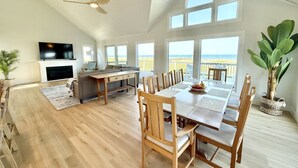 Un vaste salon doté d’un haut plafond, un coin repas avec des meubles en bois et de grandes fenêtres offrant une vue sur la plage.