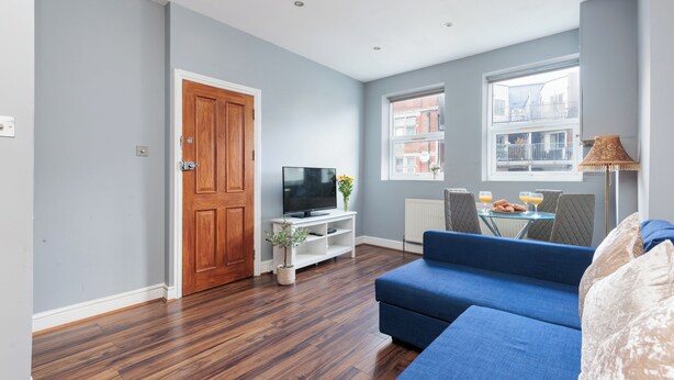 Ein Wohnzimmer mit blauem Sofa, Holzboden, einem Fernseher auf einem weißen Schrank und einem Fenster mit Blick auf Gebäude.