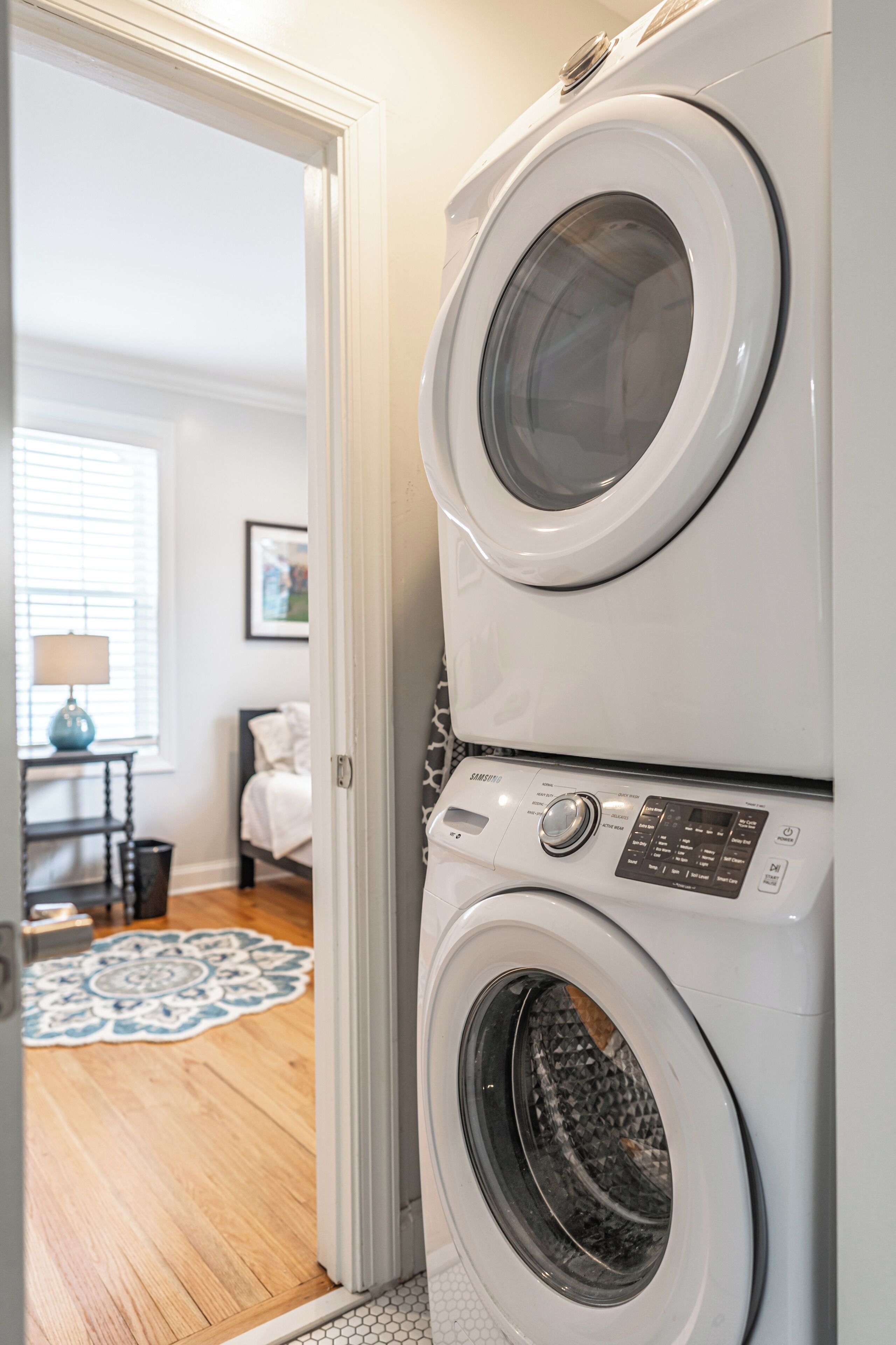A laundry area with stacked washers and dryers, a wooden floor, a patterned rug, and a bed in the background.