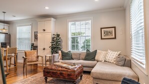 A living room with a sofa, ottoman, and chairs, a wooden coffee table, and a large window with blinds. - The McCaskill Cottage in the Village of Pinehurst!  (Pinehurst)