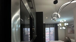 A modern hotel room with a large flat-screen TV, a bed with white bedding, a unique curved light fixture, and a view of the city through the window. - Loxwood Apartment (Accra)