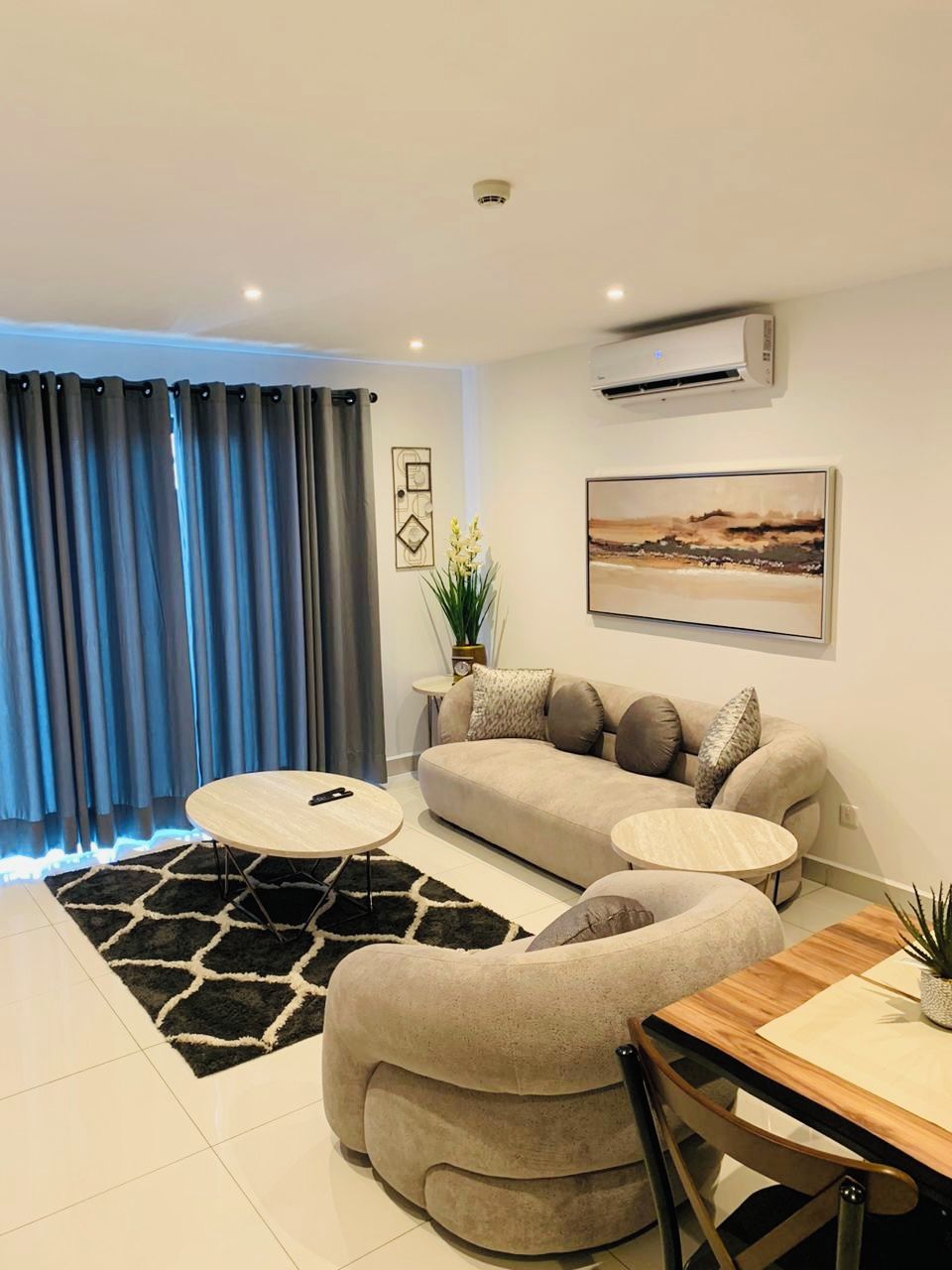 A modern living room with a sofa, two round coffee tables, a dining table with chairs, a potted plant, and a large window with curtains.
