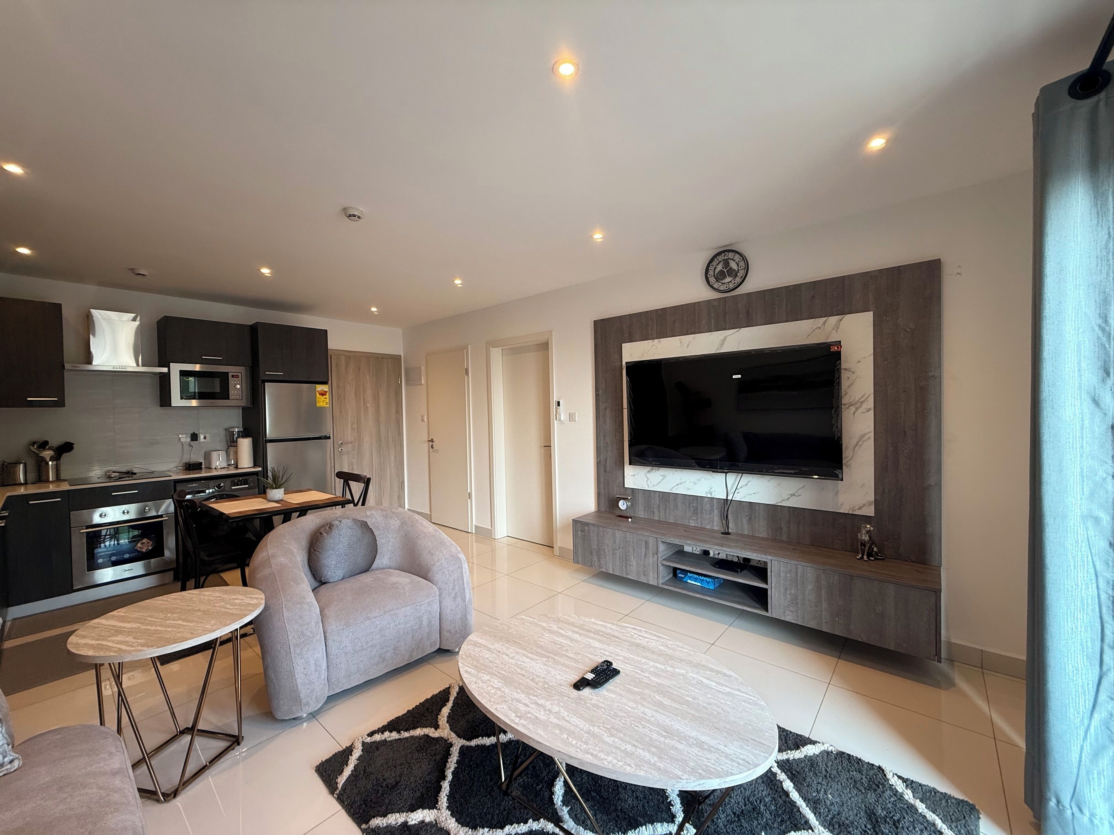 A modern kitchen with dark cabinetry, a microwave, and a built-in oven. There is a grey sofa and a small round table with a remote control on it. A large flat-screen TV is mounted on the wall above a wooden entertainment unit.