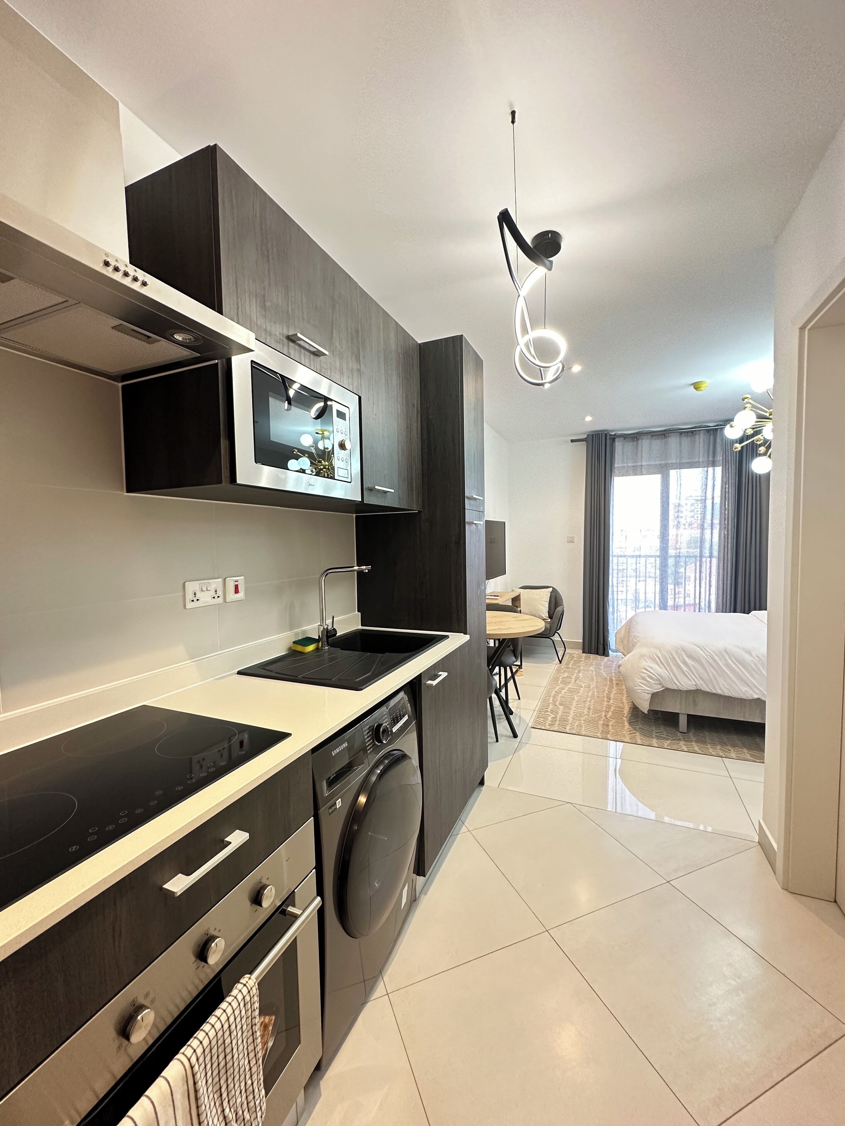 A modern kitchen with a built-in microwave, oven, and dishwasher. There is a dining area with a table and chairs, and a bed in the adjacent room.