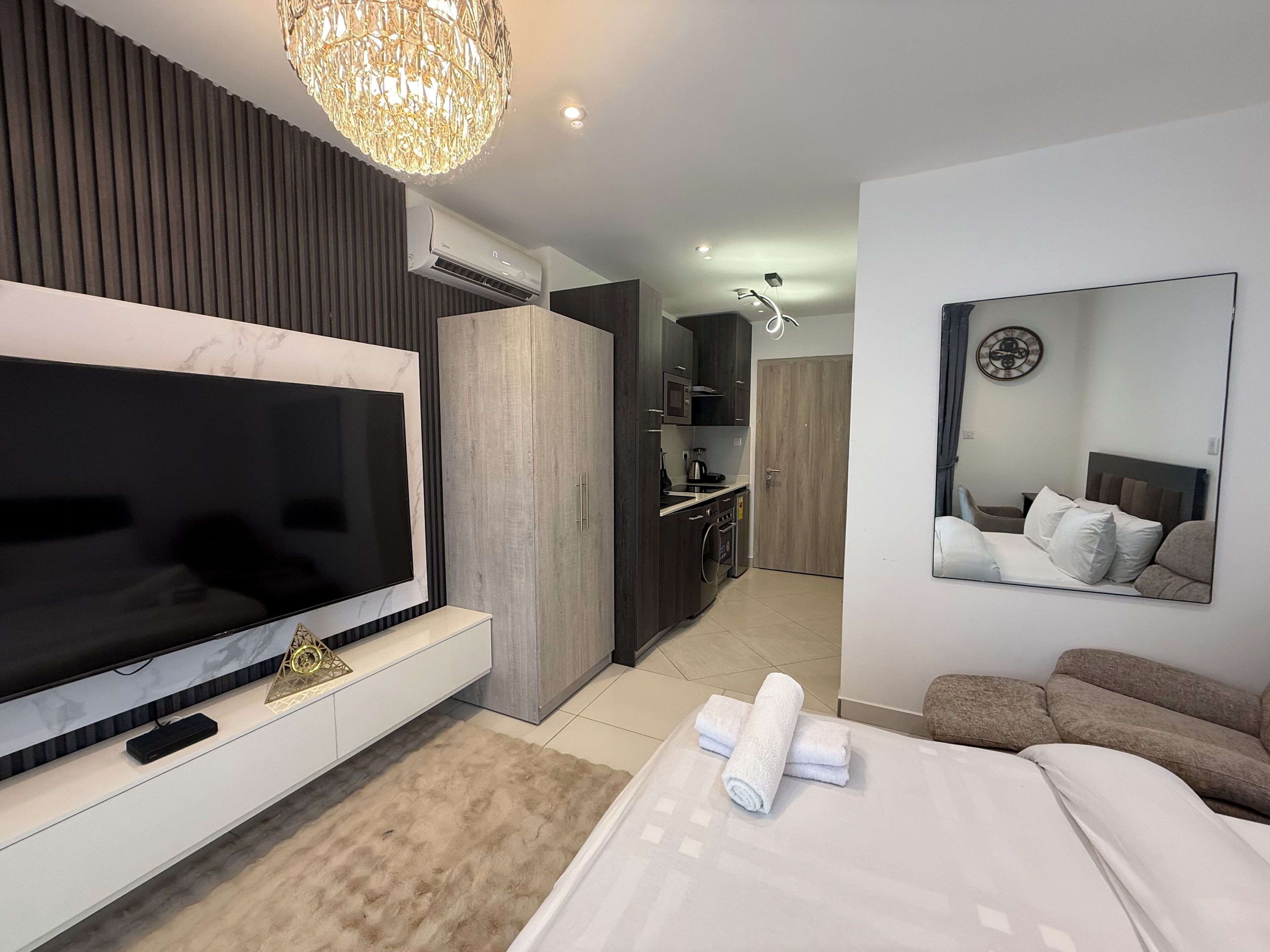 Une chambre d’hôtel moderne dotée d’une grande télévision à écran plat, d’un canapé, d’un lit avec du linge de maison blanc, d’un miroir, d’un lustre et d’une kitchenette.