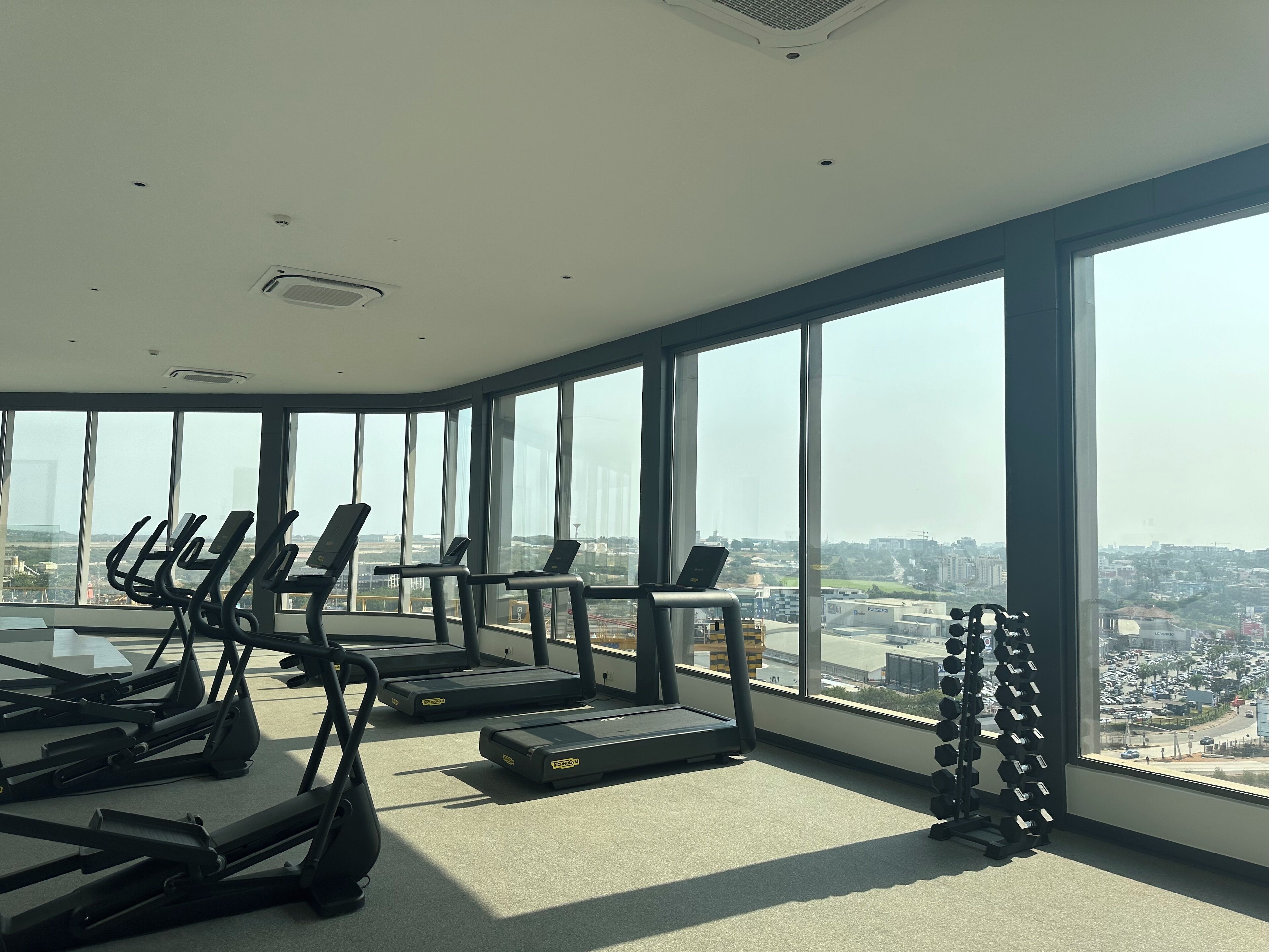 A gym with treadmills and dumbbells, large windows, and a city view.