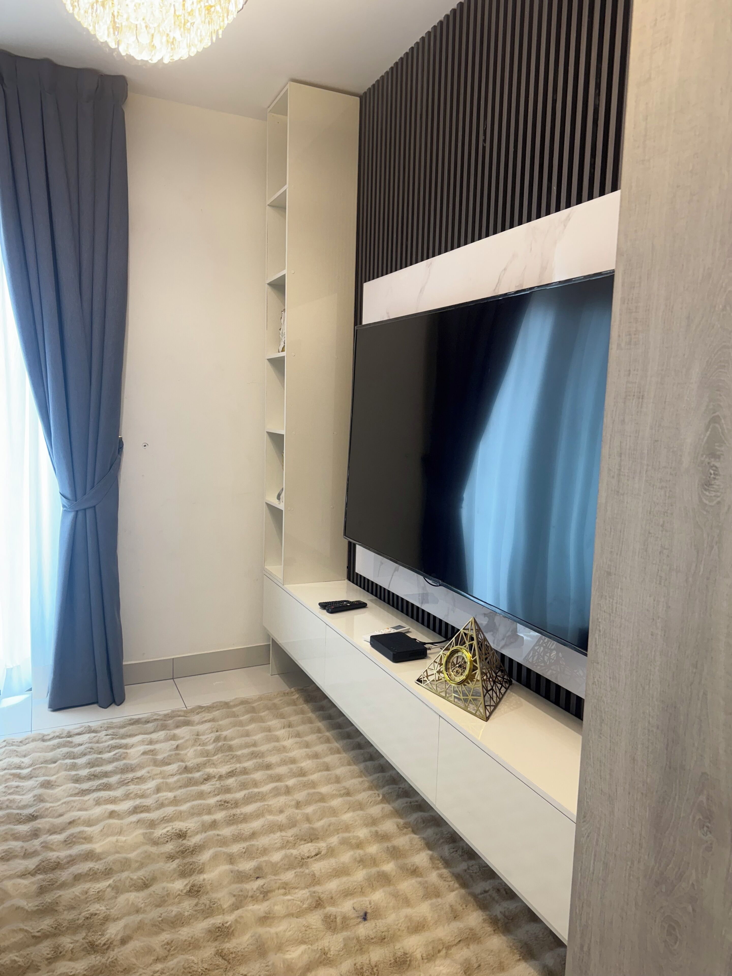 A modern living room with a large flat-screen TV mounted on a white wall, a built-in shelving unit, a fluffy beige rug, and a blue curtain.