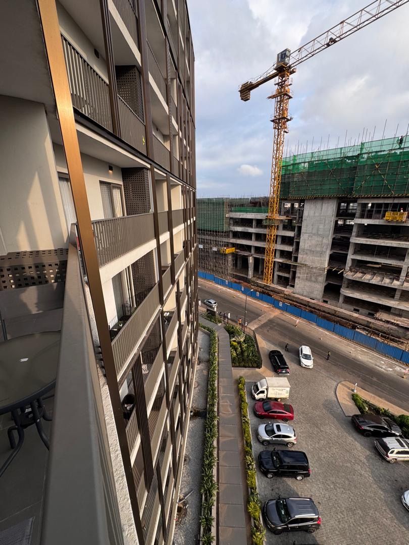 A multi-story building with balconies, a construction crane, and a parking area with cars.