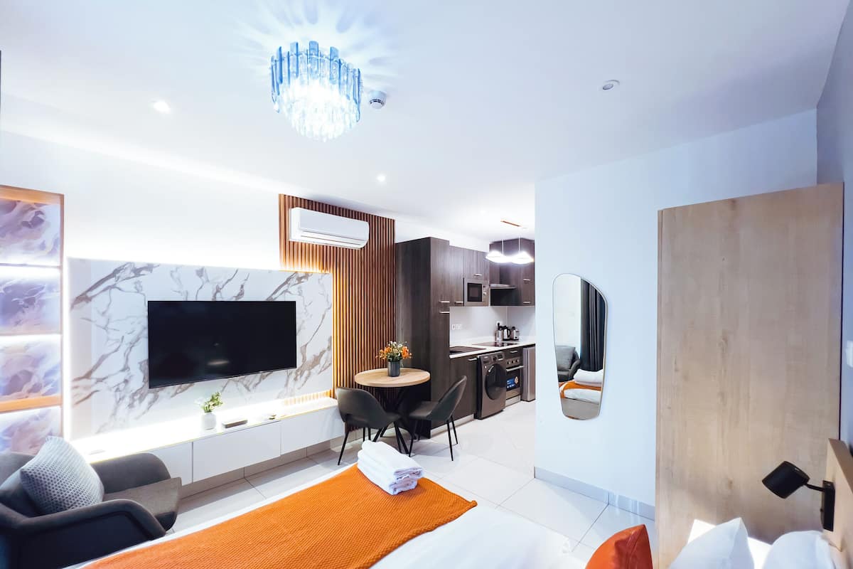 A modern hotel room with a large bed, a TV mounted on a marble wall, a dining area with a round table, and a kitchenette.