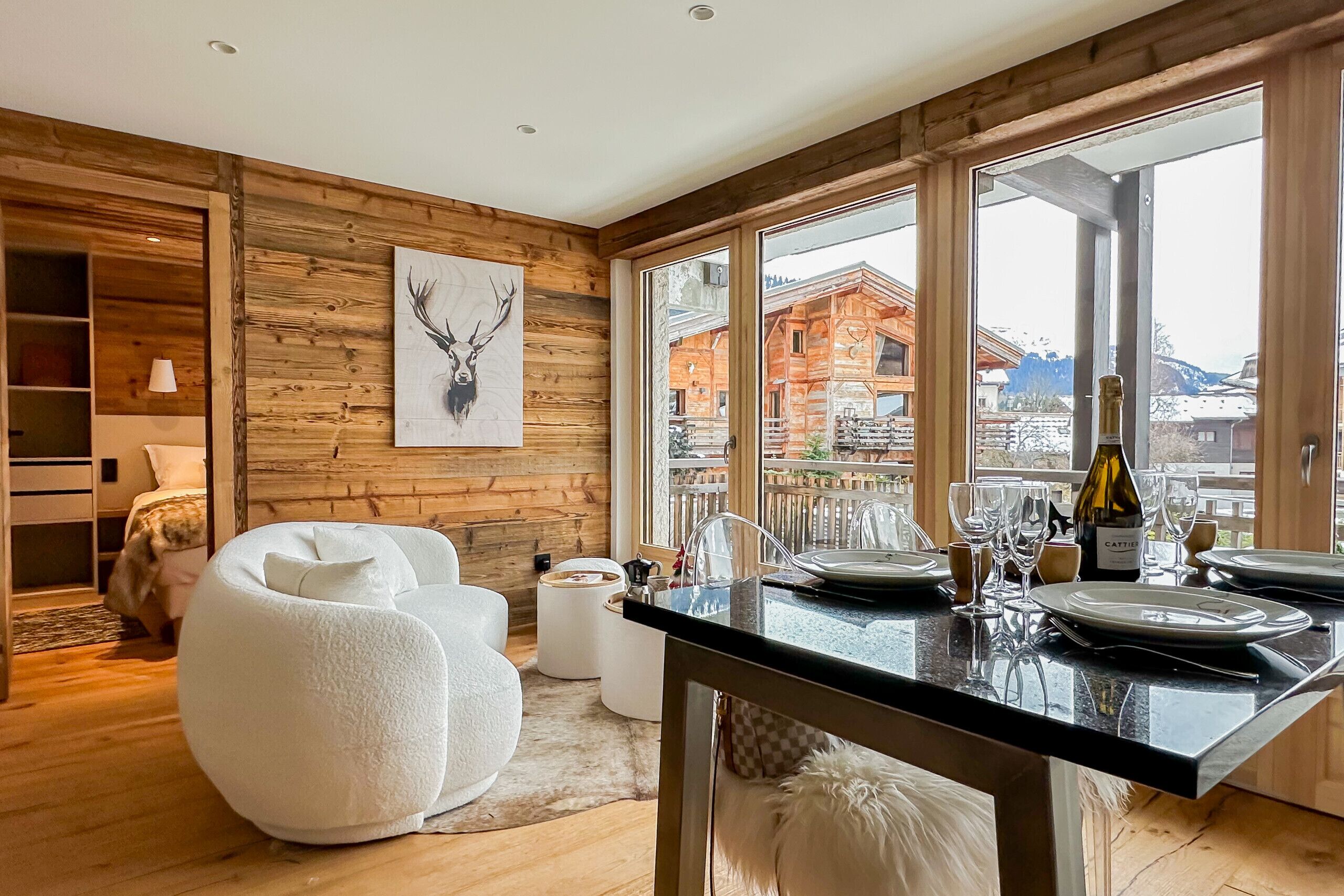 Un intérieur moderne avec un mur en bois, une décoration murale représentant une tête de cerf, un coin repas agrémenté d’une bouteille de vin Cattier, et une vue sur un bâtiment en bois à l’extérieur.