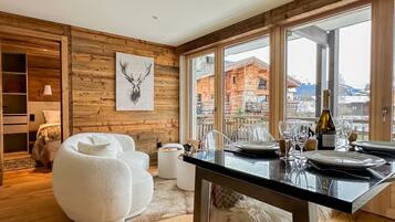 Un intérieur moderne avec un mur en bois, une décoration murale représentant une tête de cerf, un coin repas agrémenté d’une bouteille de vin Cattier, et une vue sur un bâtiment en bois à l’extérieur.
